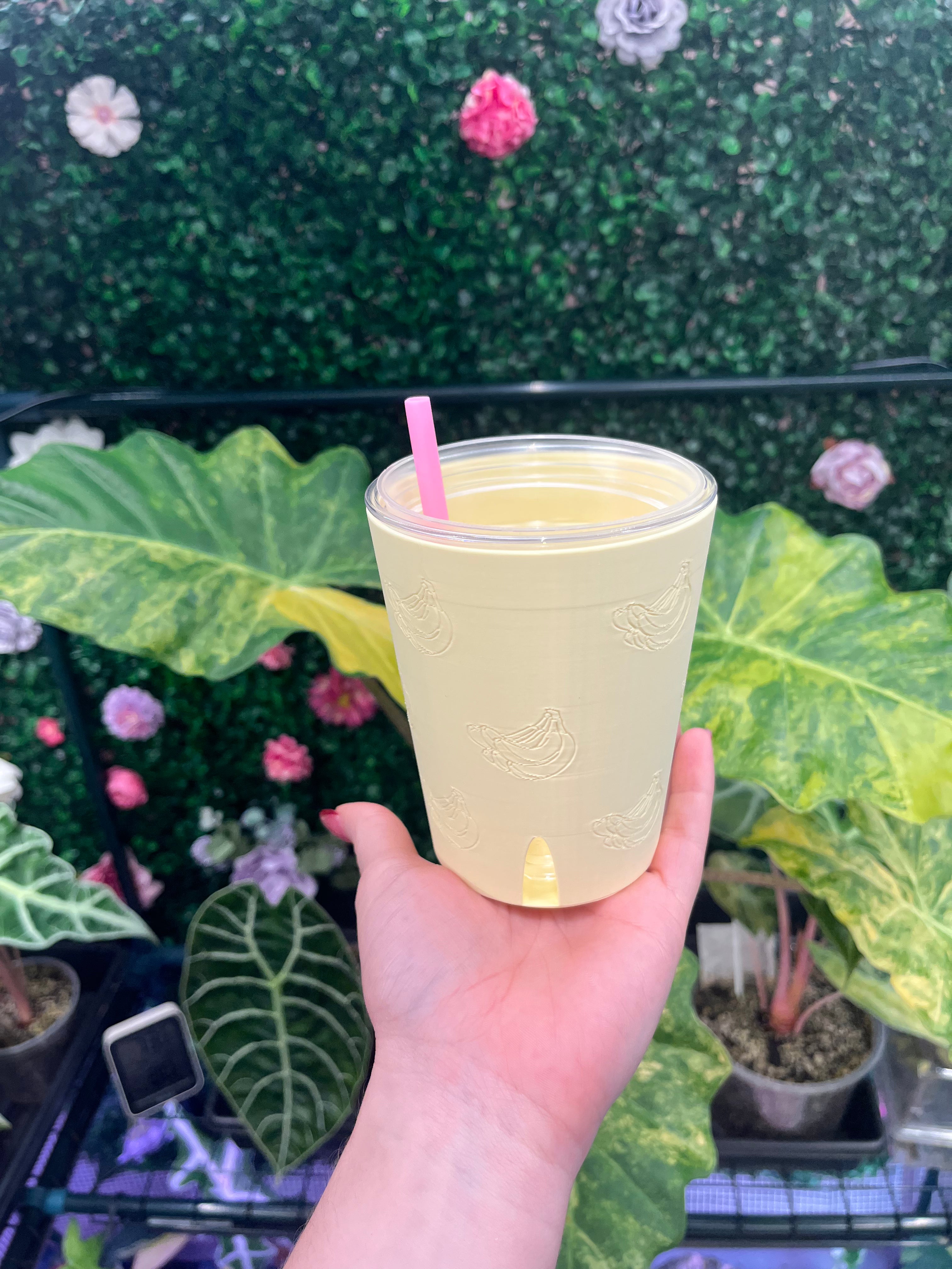A hand holding a clear plastic tumbler with a pale yellow beverage and a pink straw, set against a background of green artificial foliage and potted plants.